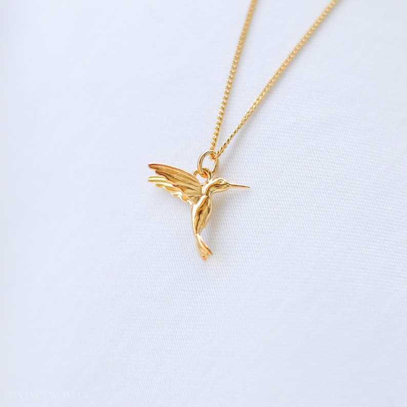 K&ouml;nnte beinhalten: Eine goldfarbene Halskette mit einem Anh&auml;nger in Form eines Kolibris. Der Kolibri befindet sich im Flug, mit ausgebreiteten Fl&uuml;geln. Die Halskette ist zierlich und ebenfalls goldfarben. Der Hintergrund ist ein weicher, wei&szlig;er Stoff.