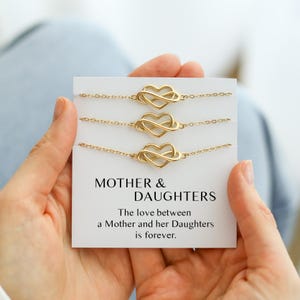 May include: Three gold-colored bracelets with heart and infinity symbol charms are displayed on a white card. The card reads "MOTHER & DAUGHTERS" with a message about the love between a mother and daughters.