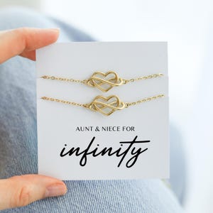 May include: Two gold-colored bracelets with heart-shaped infinity symbols are displayed on a white card. The card reads "AUNT & NIECE FOR infinity" in black script. The bracelets are on a delicate chain.