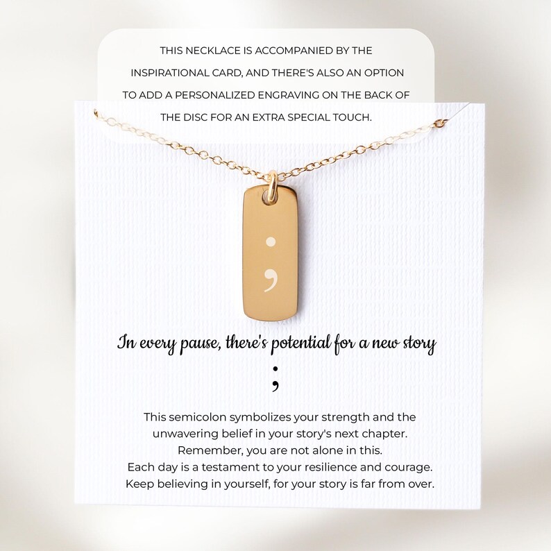 Puede incluir: Un collar con colgante rectangular dorado grabado con un punto y coma, presentado en una tarjeta blanca con el texto: In every pause, there's potential for a new story. La tarjeta incluye texto adicional sobre el significado del s&iacute;mbolo.