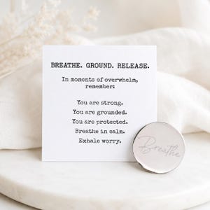 Peut inclure: Une carte blanche avec le texte "BREATHE. GROUND. RELEASE." et des phrases inspirantes. Une petite pièce ronde argentée avec le mot "Breathe" gravé est à côté de la carte. Les articles sont sur une surface en marbre blanc.
