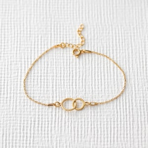 Puede incluir: Una delicada pulsera dorada con dos c&iacute;rculos entrelazados como elemento central. La pulsera tiene una fina cadena y un cierre ajustable. La joya est&aacute; sobre un fondo blanco texturizado.