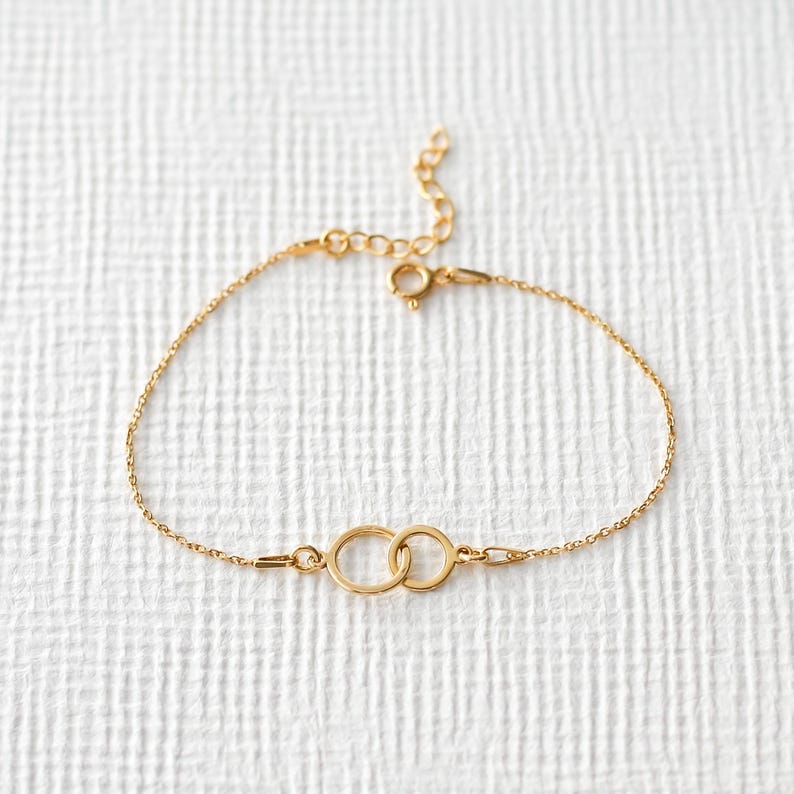 Puede incluir: Una delicada pulsera dorada con dos c&iacute;rculos entrelazados como elemento central. La pulsera tiene una fina cadena y un cierre ajustable. La joya est&aacute; sobre un fondo blanco texturizado.
