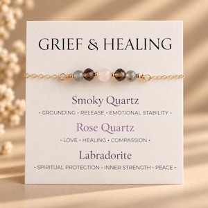 Könnte beinhalten: Ein zartes Goldkettenarmband mit einer zentralen Anordnung aus fünf Perlen: zwei grauen Labradoriten, zwei dunkelbraunen Rauchquarzen und einem blassrosa Rosenquarz. Das Armband ist auf einer weißen Karte mit der Aufschrift GRIEF & HEALING.