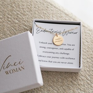 May include: A gold-colored necklace with a round pendant that reads "endo warrior" in a white box with the text "Endometriosis Warrior". The box is open, revealing the necklace and a card with inspirational text. The box is white with the text "Vinci WOMAN".
