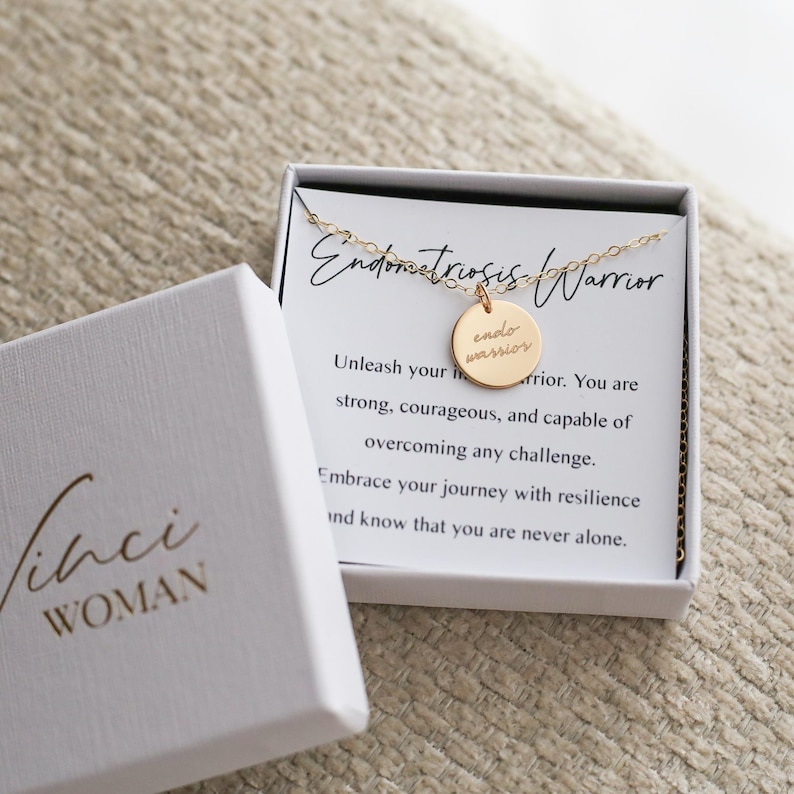 May include: A gold-colored necklace with a round pendant that reads "endo warrior" in a white box with the text "Endometriosis Warrior". The box is open, revealing the necklace and a card with inspirational text. The box is white with the text "Vinci WOMAN".