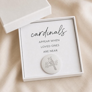 Può includere: Una scatola regalo bianca con una carta che recita "cardinals appear when loved ones are near". Una moneta d'argento con un disegno di uccello cardinale è al centro della carta. La scatola è aperta, rivelando il contenuto.