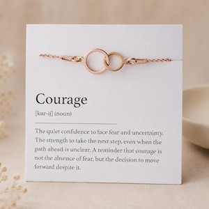 Pode incluir: Uma pulseira de ouro rosa com dois anéis circulares entrelaçados presos a uma corrente delicada, apresentada num cartão branco impresso com a palavra Courage, a sua transcrição fonética e uma definição do substantivo em texto preto.
