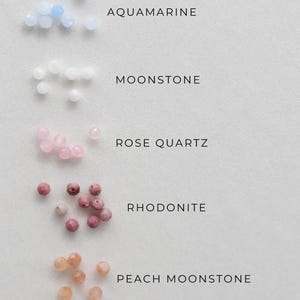 May include: An overhead shot displays five types of gemstone beads: aquamarine, moonstone, rose quartz, rhodonite, and peach moonstone. Each gemstone is accompanied by its name in a clean, sans-serif font. The beads are small and round, with varying shades of blue, white, pink, and peach.