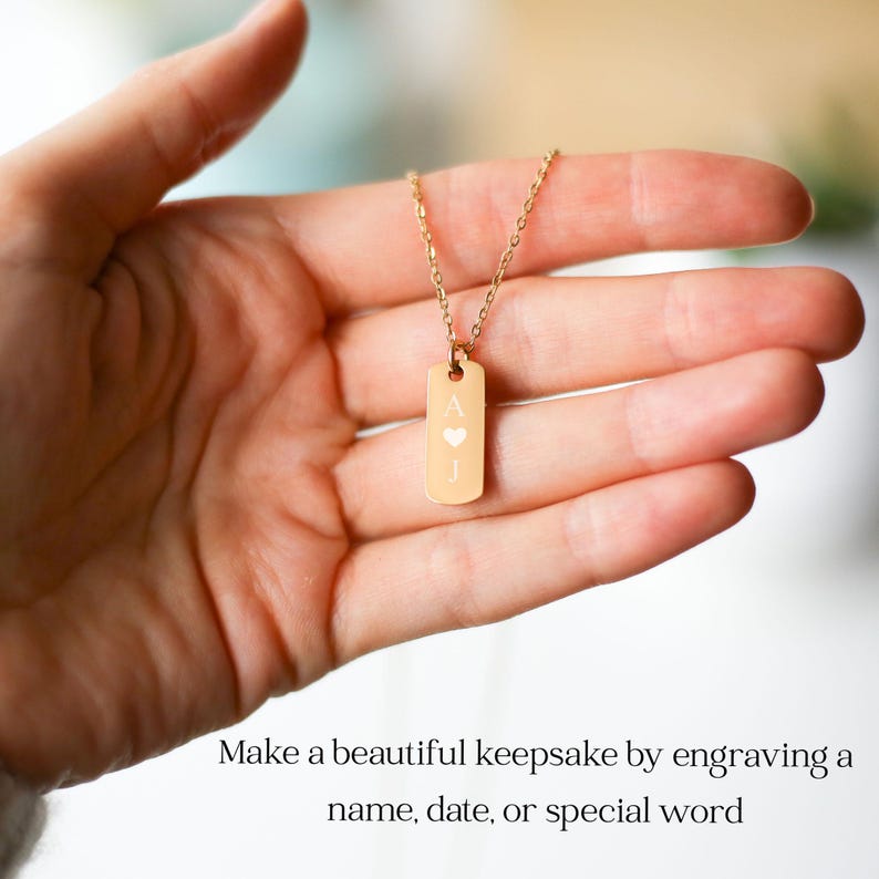 Puede incluir: Un collar con colgante rectangular de tono dorado que presenta la letra A grabada, un peque&ntilde;o s&iacute;mbolo de coraz&oacute;n y la letra J. El colgante cuelga de una delicada cadena dorada. El texto inferior dice: Make a beautiful keepsake by engraving a name, date, or special word.