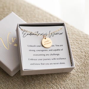 May include: A gold-colored necklace with a round pendant that reads "endo warrior" in a script font. The necklace is displayed in a white box with the words "Endometriosis Warrior" on a card. The box is open, revealing the necklace.