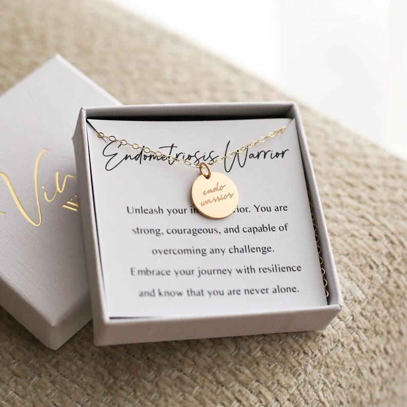 May include: A gold-colored necklace with a round pendant that reads "endo warrior" in a script font. The necklace is displayed in a white box with the words "Endometriosis Warrior" on a card. The box is open, revealing the necklace.