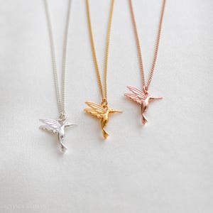 K&ouml;nnte beinhalten: Drei Halsketten mit Anh&auml;ngern in Form von Kolibris in Silber, Gold und Ros&eacute;gold. Jede Halskette hat einen detaillierten Kolibri-Anh&auml;nger, der an einer zarten Kette h&auml;ngt. Die Anh&auml;nger werden vor einem wei&szlig;en Hintergrund pr&auml;sentiert.