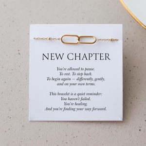 Könnte beinhalten: Ein goldfarbenes Armband mit einem verbundenen Design wird auf einer weißen Karte präsentiert. Die Karte trägt die Aufschrift "NEW CHAPTER" mit inspirierendem Text. Das Armband ist ein zartes Schmuckstück, ideal zum Verschenken oder Tragen.