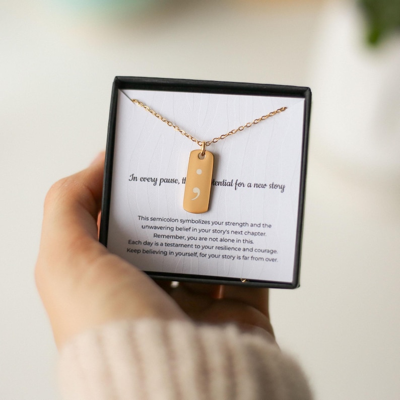 Puede incluir: Un collar de tono dorado con un colgante rectangular que presenta un s&iacute;mbolo de punto y coma. El colgante se muestra sobre una tarjeta blanca dentro de una caja negra, con el texto: In every pause, the potential for a new story.