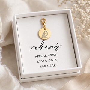 Puede incluir: Un charm dorado con un grabado de pájaro, sujeto a un cierre, presentado en una caja blanca. La caja tiene la palabra "robins" escrita en cursiva, con la frase "appear when loved ones are near" debajo.