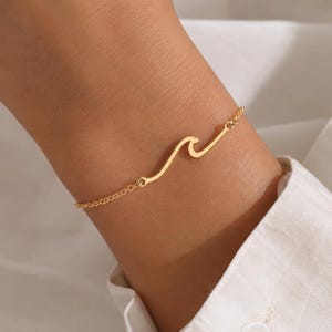 Puede incluir: Un brazalete dorado con un diseño de ola. El brazalete está hecho de una cadena delicada con un colgante en forma de ola. La joya se lleva en la muñeca. El fondo es blanco.