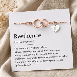 Peut inclure: Bracelet en or rose avec deux cercles entrelacés et un charm en forme de cœur gravé de la lettre "R". Le bracelet est présenté sur une carte avec la définition de "Résilience".