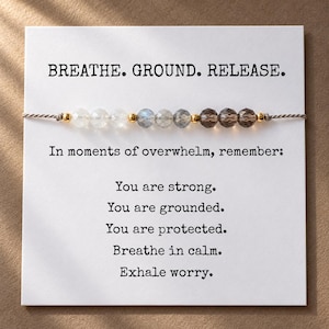 Könnte beinhalten: Ein Perlenarmband auf einer weißen Karte mit dem Text BREATHE. GROUND. RELEASE. In moments of overwhelm, remember: You are strong. You are grounded. You are protected. Breathe in calm. Exhale worry. Das Armband besteht aus facettierten Perlen in Weiß, Grau und Braun.