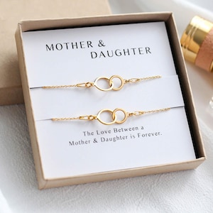 May include: A gold bracelet set for a mother and daughter, presented in a gift box. Each delicate chain features two interlocking circles. The card inside reads "Mother & Daughter" and "The Love Between a Mother & Daughter is Forever."