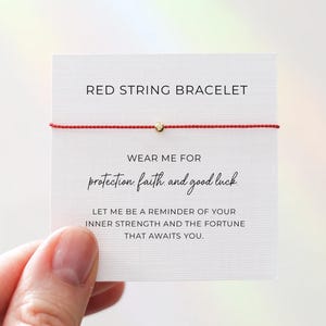 Könnte beinhalten: Ein rotes Armband mit einer goldenen Perle, präsentiert auf einer weißen Karte. Auf der Karte steht "RED STRING BRACELET" und ein Text über Schutz, Glauben und Glück. Das Armband wird von einer Hand gehalten.