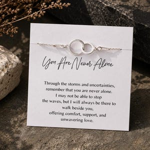 May include: A silver bracelet featuring two linked circles, presented on a white card. The card displays the text "You Are Never Alone" in cursive, accompanied by an encouraging message. The bracelet is on a stone surface.