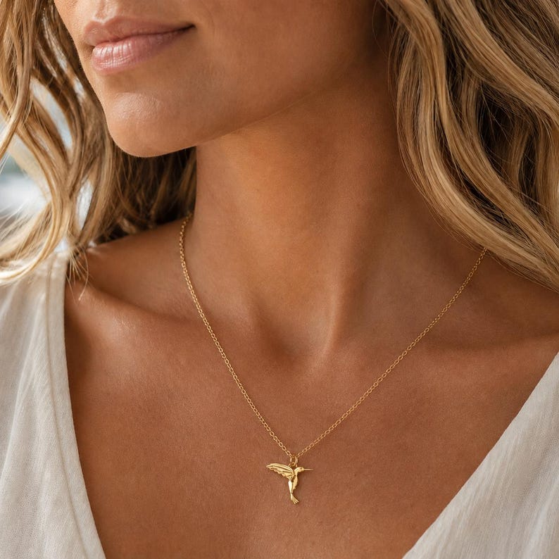 May include: A gold-colored necklace featuring a hummingbird pendant. The pendant is intricately detailed, showcasing the bird's wings and body. The necklace is worn around the neck, with the pendant resting on the chest. The chain is delicate and appears to be made of gold.