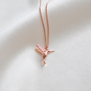 K&ouml;nnte beinhalten: Halskette mit Anh&auml;nger in Form eines Kolibris aus Ros&eacute;gold. Der filigrane Anh&auml;nger zeigt einen detaillierten Kolibri mit ausgebreiteten Fl&uuml;geln, der an einer passenden Kette h&auml;ngt. Die Halskette wird auf einem wei&szlig;en, strukturierten Hintergrund pr&auml;sentiert.