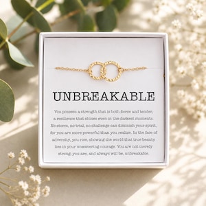 May include: A gold bracelet with two linked circles, displayed in a white gift box. The box has the word "UNBREAKABLE" and motivational text. The bracelet is set against a white backdrop with floral decorations.