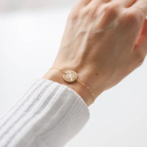 Könnte beinhalten: Ein zartes, goldfarbenes Armband mit einem kleinen, runden Anhänger mit einem detaillierten Blumendesign. Das Armband wird am Handgelenk getragen und zeigt seine feine Kette und den gravierten Anhänger. Die Ästhetik ist minimalistisch und elegant.