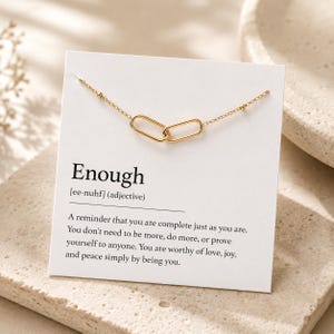 May include: A gold-toned chain necklace featuring two interlocking oval links, displayed on a white card. The card includes the printed text "Enough [ee-nuhf] (adjective)" followed by a paragraph describing the word as a reminder of self-worth.