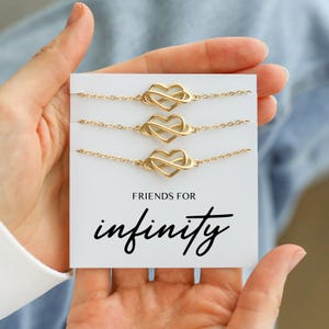 Könnte beinhalten: Drei goldfarbene Infinity-Herz-Armbänder auf einer weißen Karte. Auf der Karte steht "FRIENDS FOR infinity" in schwarzer Schrift. Die Armbänder werden auf einer weißen Karte präsentiert, die von einer Person gehalten wird.