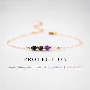 Puede incluir: Una delicada pulsera de cadena dorada con cuatro cuentas de piedras preciosas: turmalina negra, ojo de tigre, amatista y cuarzo rosa. La palabra "PROTECTION" está impresa debajo, con los nombres de las piedras.