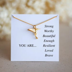May include: A gold-colored hummingbird pendant necklace on a white card. The card reads "YOU ARE..." followed by "Strong, Worthy, Beautiful, Enough, Resilient, Loved, Brave."