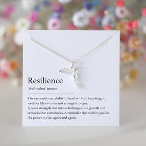 K&ouml;nnte beinhalten: Eine silberne Halskette mit einem Anh&auml;nger in Form eines Kolibris auf einer wei&szlig;en Karte. Auf der Karte steht "Resilienz" mit einer Definition. Die Halskette ist zart und der Kolibri ist im Flug. Der Hintergrund ist verschwommen mit bunten Blumen.