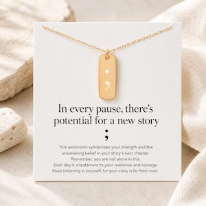 Puede incluir: Un collar de tono dorado con un colgante rectangular grabado con un s&iacute;mbolo de punto y coma. El colgante se muestra sobre una tarjeta blanca impresa con el texto: In every pause, there's potential for a new story, seguido de un punto y coma.