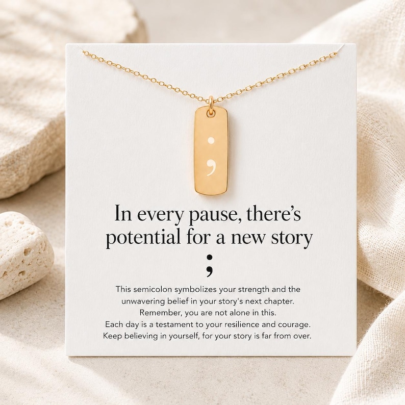 Puede incluir: Un collar de tono dorado con un colgante rectangular grabado con un s&iacute;mbolo de punto y coma. El colgante se muestra sobre una tarjeta blanca impresa con el texto: In every pause, there's potential for a new story, seguido de un punto y coma.