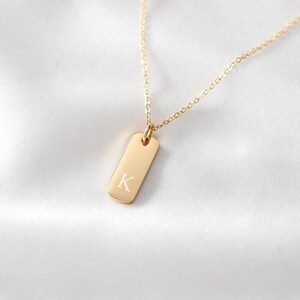 Puede incluir: Un collar de tono dorado con un colgante rectangular de esquinas redondeadas. El colgante est&aacute; grabado con la letra may&uacute;scula K en una fuente con serifa. El colgante cuelga de una cadena fina y delicada de tono dorado con eslabones uniformes.