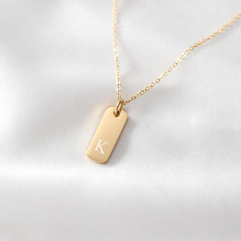 Puede incluir: Un collar de tono dorado con un colgante rectangular de esquinas redondeadas. El colgante est&aacute; grabado con la letra may&uacute;scula K en una fuente con serifa. El colgante cuelga de una cadena fina y delicada de tono dorado con eslabones uniformes.