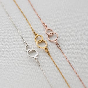 Puede incluir: Tres delicadas pulseras de cadena en plata, oro y oro rosa. Cada pulsera presenta una serie de c&iacute;rculos interconectados como elemento central del dise&ntilde;o. Las pulseras se exhiben sobre una superficie blanca texturizada.