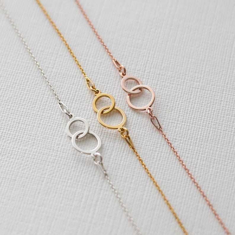 Puede incluir: Tres delicadas pulseras de cadena en plata, oro y oro rosa. Cada pulsera presenta una serie de c&iacute;rculos interconectados como elemento central del dise&ntilde;o. Las pulseras se exhiben sobre una superficie blanca texturizada.