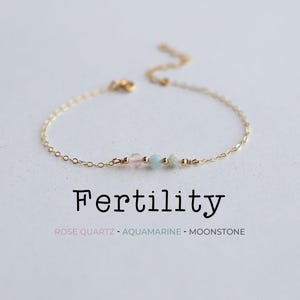 May include: Delicate gold chain bracelet with three gemstone beads: pink rose quartz, light blue aquamarine, and white moonstone. The word "Fertility" is written below the bracelet.