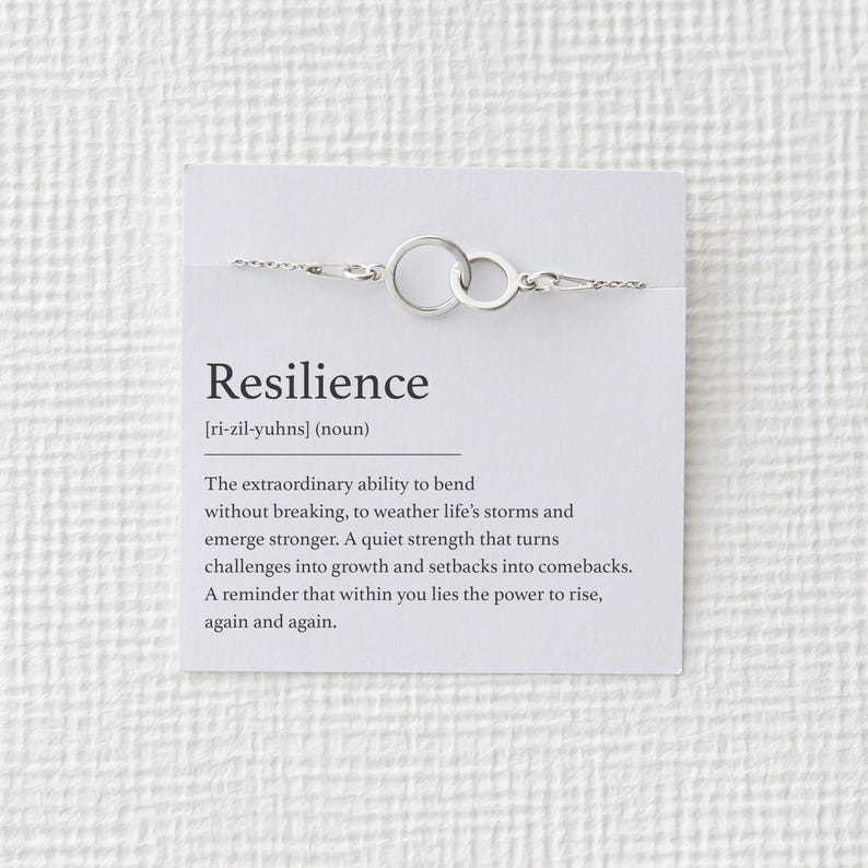 Puede incluir: Un brazalete plateado con dos c&iacute;rculos entrelazados, exhibido en una tarjeta blanca. La tarjeta presenta la palabra "Resiliencia" y su definici&oacute;n, enfatizando la fuerza interior. Una delicada pieza de joyer&iacute;a.