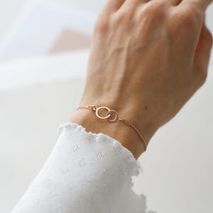 Puede incluir: Pulsera de oro rosa con dos c&iacute;rculos entrelazados. La fina pulsera de cadena se muestra en una mu&ntilde;eca, con una manga blanca con volantes en el fondo. Los c&iacute;rculos son un elemento de dise&ntilde;o clave, que simboliza la conexi&oacute;n y la unidad.