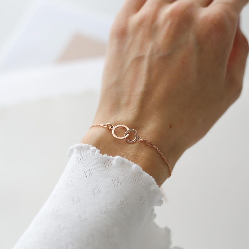 Puede incluir: Pulsera de oro rosa con dos c&iacute;rculos entrelazados. La fina pulsera de cadena se muestra en una mu&ntilde;eca, con una manga blanca con volantes en el fondo. Los c&iacute;rculos son un elemento de dise&ntilde;o clave, que simboliza la conexi&oacute;n y la unidad.