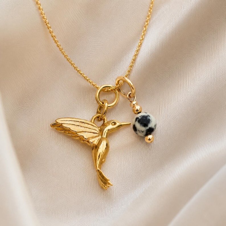 May include: Gold-colored necklace featuring a hummingbird pendant with outstretched wings and a small, round charm with black and white spots. The necklace hangs on a delicate chain, set against a soft, white background.