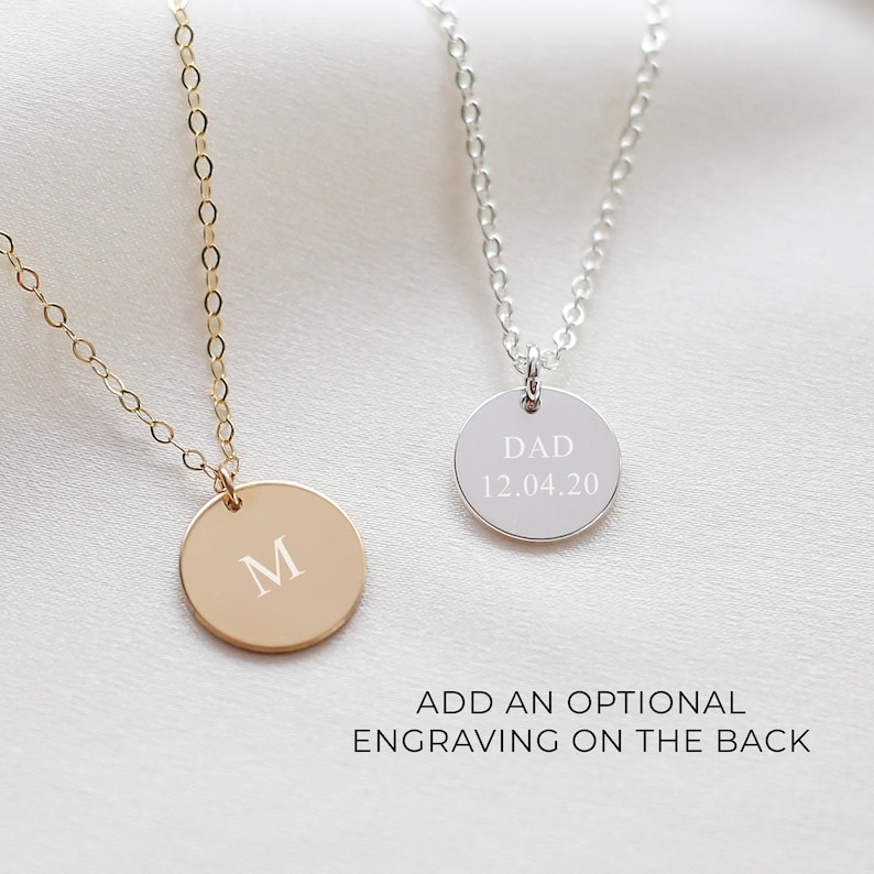 May include: Two delicate necklaces with round pendants. One is gold-toned with the letter "M" engraved, the other is silver-toned with "DAD 12.04.20" engraved. The text "ADD AN OPTIONAL ENGRAVING ON THE BACK" is below the necklaces.