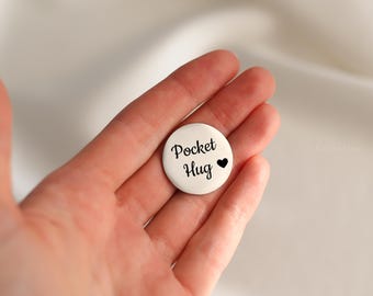 May include: A white, round pocket hug token with the words "Pocket Hug" in black script and a black heart symbol. The token is held in a hand against a soft, neutral background.
