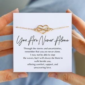 Könnte beinhalten: Ein goldfarbenes Kettenarmband mit einem zentralen Unendlichkeits-Herz-Anhänger auf einer weißen Karte. Die Karte zeigt den Text "You Are Never Alone" sowie eine gedruckte Botschaft über Unterstützung, Trost und beständige Liebe.