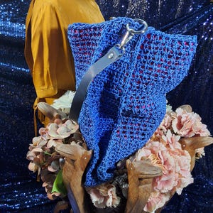 May include: A vibrant blue crocheted handbag with a red interior and a gray leather strap. The bag features a silver clasp and is displayed with a bouquet of pink and white flowers and a mustard-colored garment.